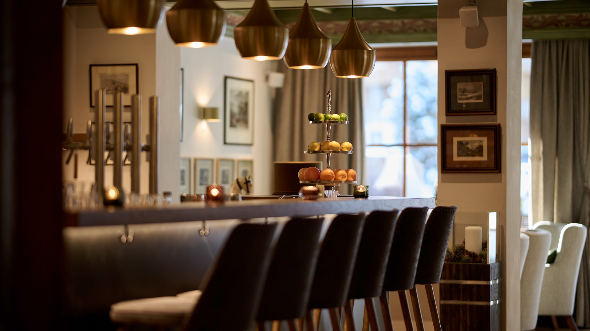 Elegante Hotelbar mit hohen Stühlen, goldenen Pendelleuchten und Obstetagere im warmen Licht.
