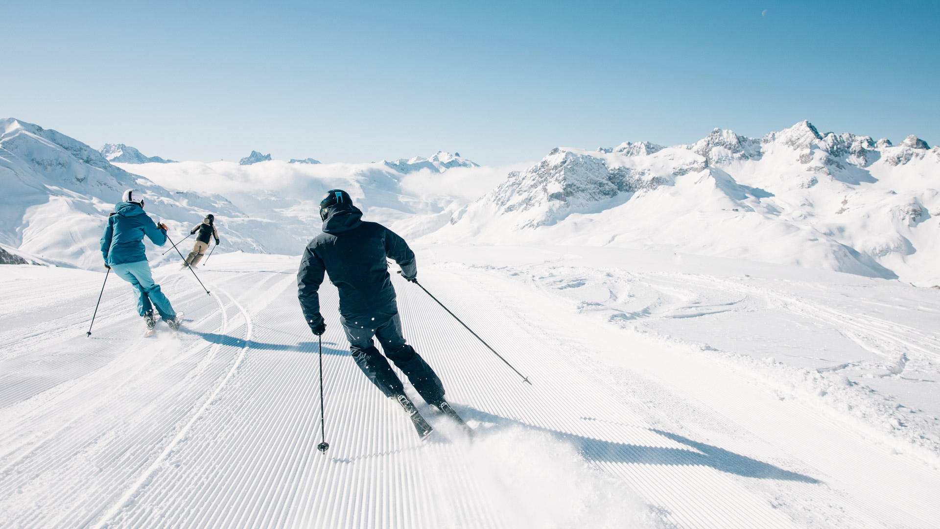 Drei Skifahrere auf einer perfekt präparierten Skipiste bei strahlenden Sonnenschein und blauem Himmel.